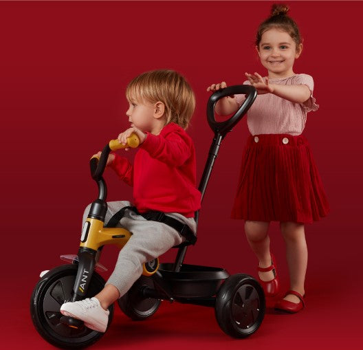 Two children on a red background, one on a tricycle and the other standing.